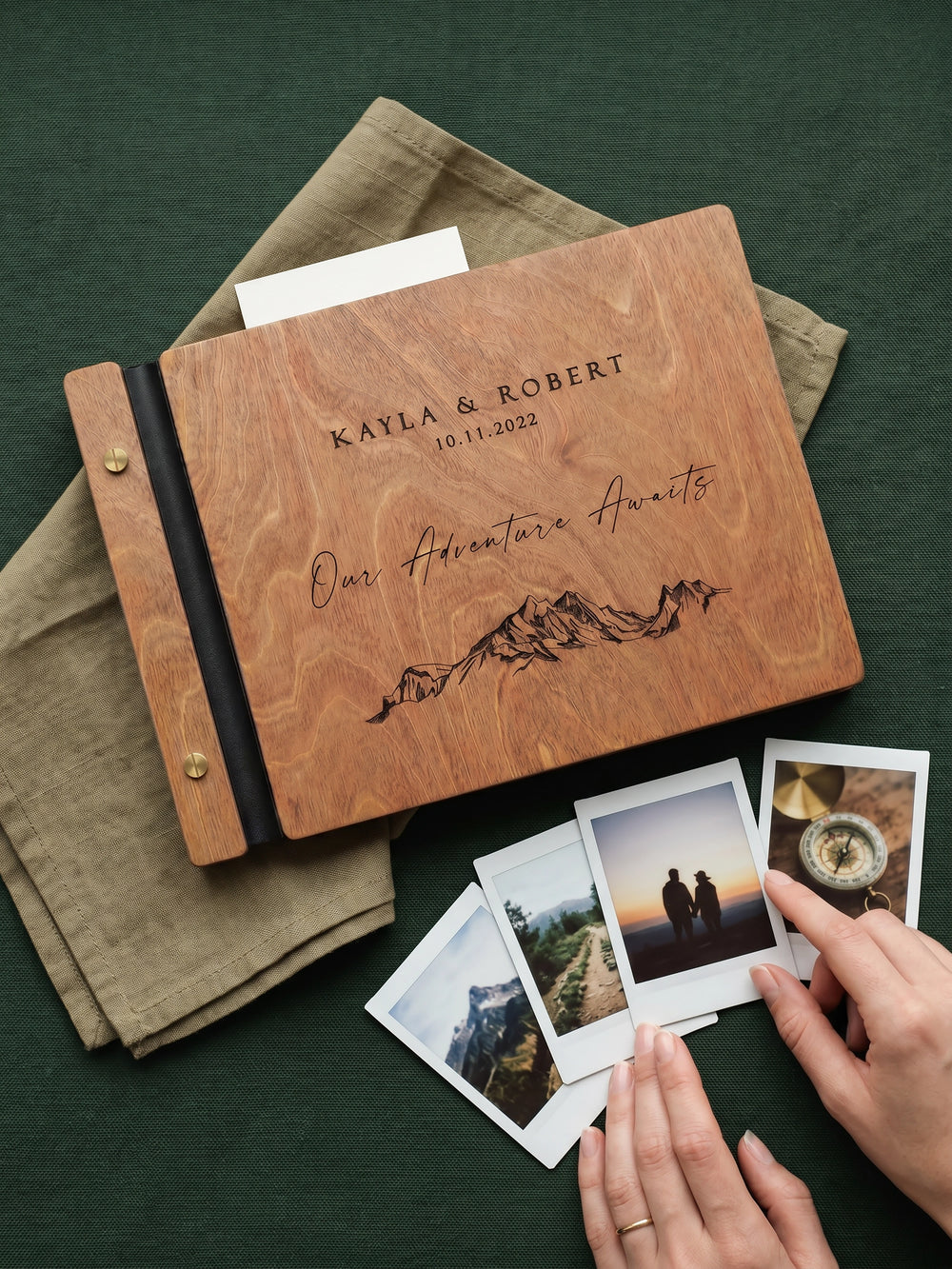 Hazelnut wood Our Adventure Awaits photo guest book engraved Kayla & Robert 10.11.2022 with a mountain range, on a green cloth with instant photos held in a hand
