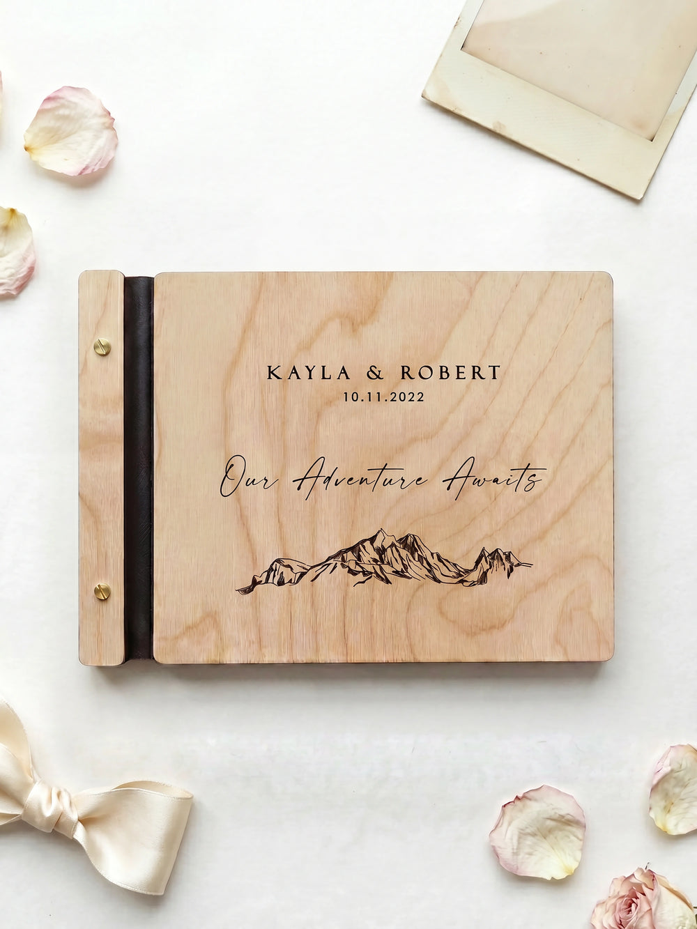Natural wood Our Adventure Awaits photo guest book with brass screws, laid on a white cloth with rose petals, a ribbon and an instant photo frame