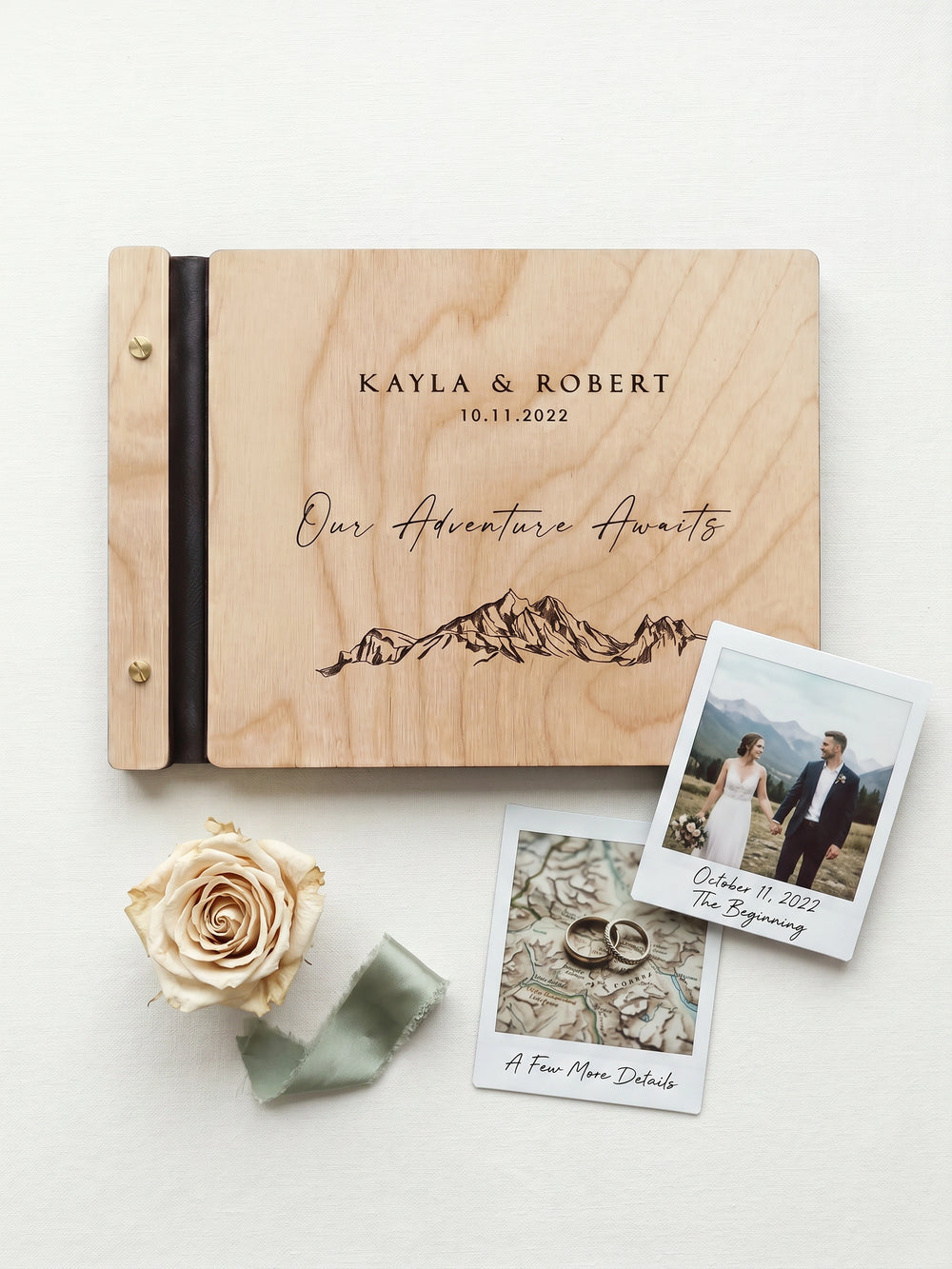 Natural wood Our Adventure Awaits photo guest book on a white cloth with a dried rose, a sage ribbon and two instant photos