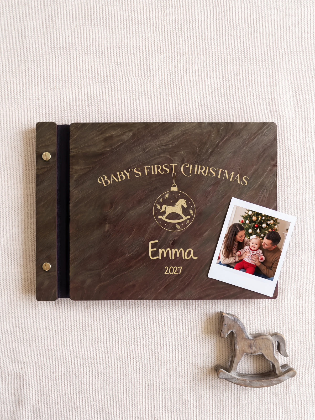 Jacobean wood Baby's First Christmas photo book lying flat on ivory knit fabric, the cover engraved with “BABY'S FIRST CHRISTMAS”, “Emma” and “2027”; two gold-tone screws along the black vegan leather spine