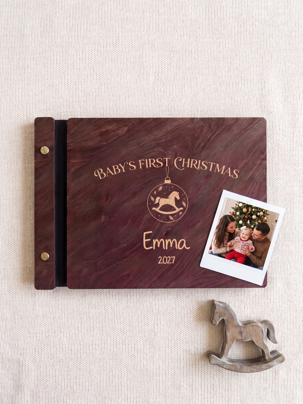 Jacobean wood Baby's First Christmas photo book lying flat on ivory knit fabric, the cover engraved with “BABY'S FIRST CHRISTMAS”, “Emma” and “2027”; two gold-tone screws along the black vegan leather spine