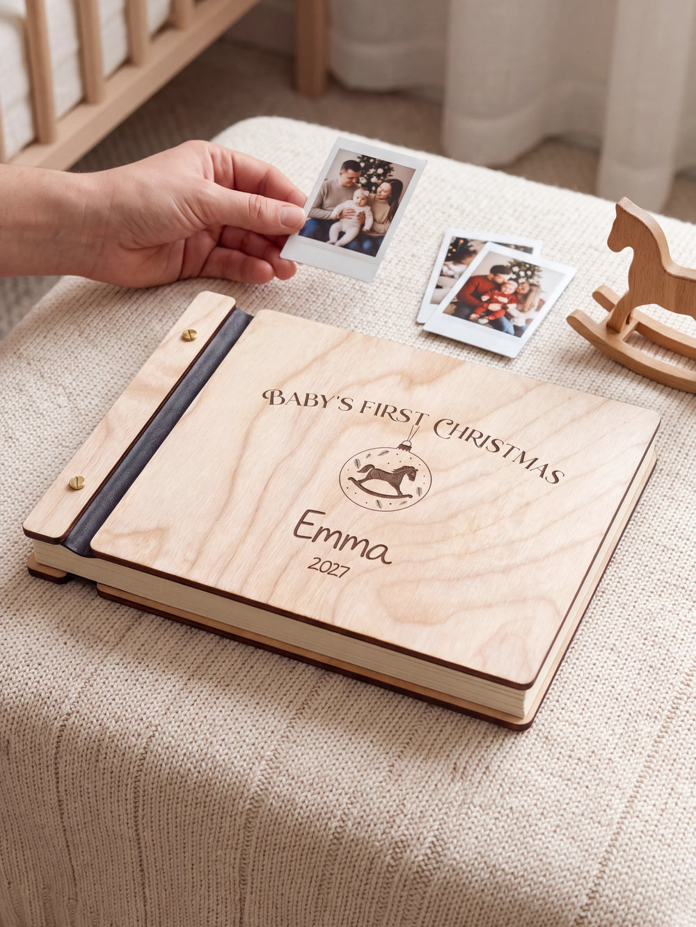 Natural wood Baby's First Christmas photo book on a light table while a hand picks an instant photo to add; the engraved cover reads “BABY'S FIRST CHRISTMAS”, “Emma” and “2027”