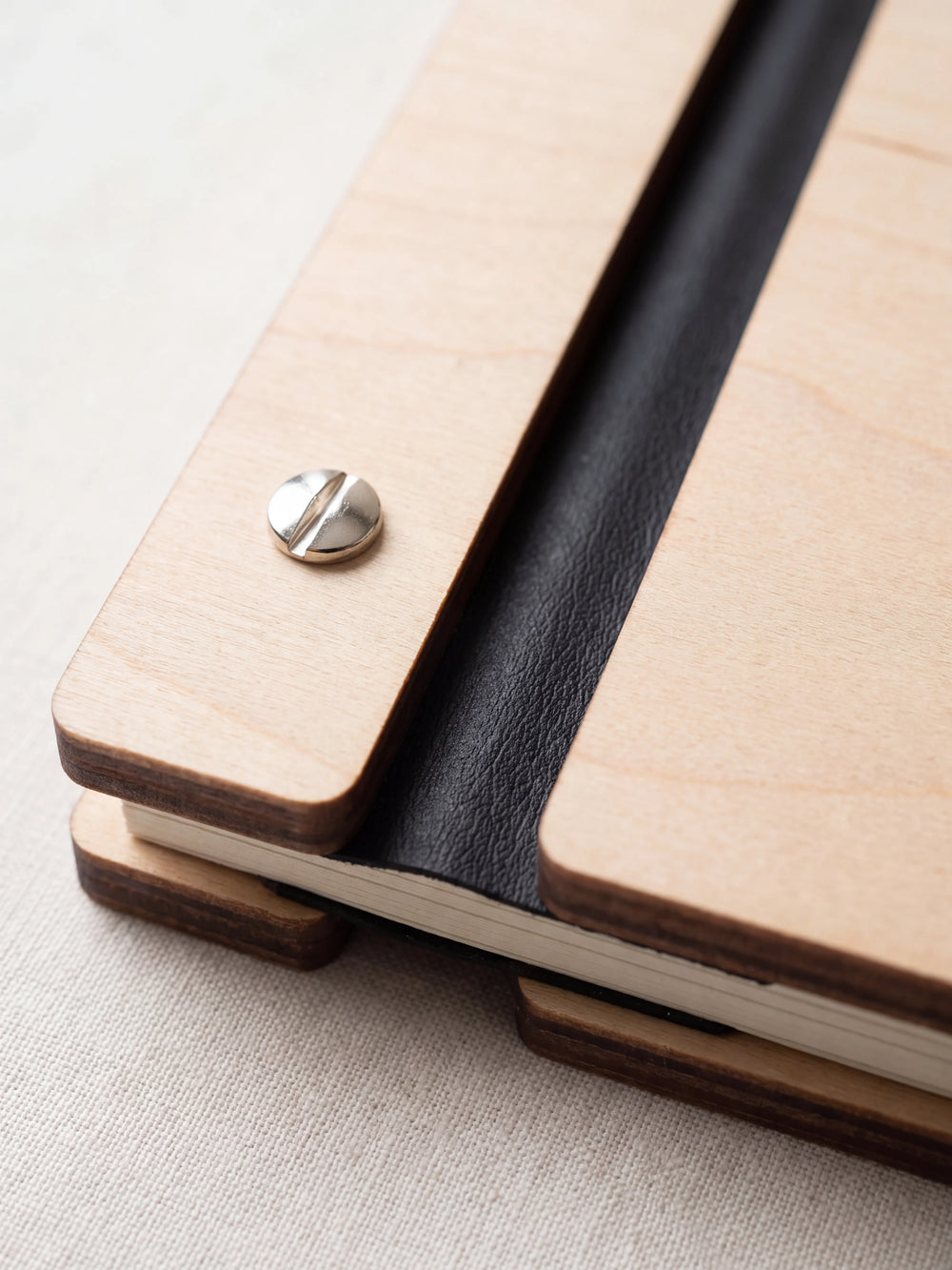 Close-up of a polished silver screw in the Natural wood strip beside the black vegan leather binding, the cream sheet stack visible below