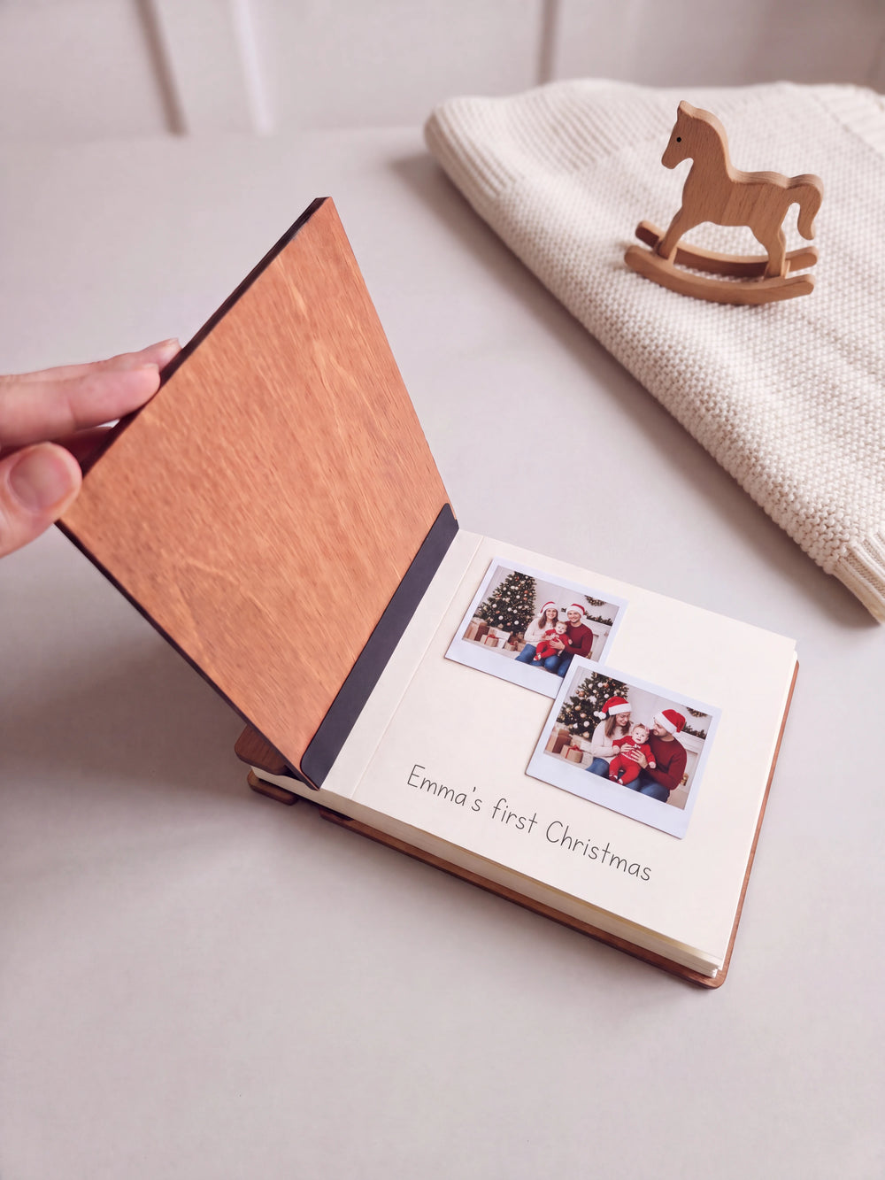 The Hazelnut Baby's First Christmas photo book open on a table with instant photos and a short handwritten note on a cream page; the black hinge strip is visible