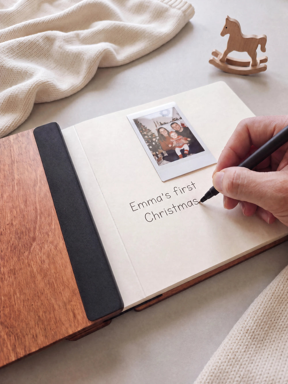 A hand writing a note in fine black pen beneath an instant photo on the cream page of the open Hazelnut Baby's First Christmas photo book