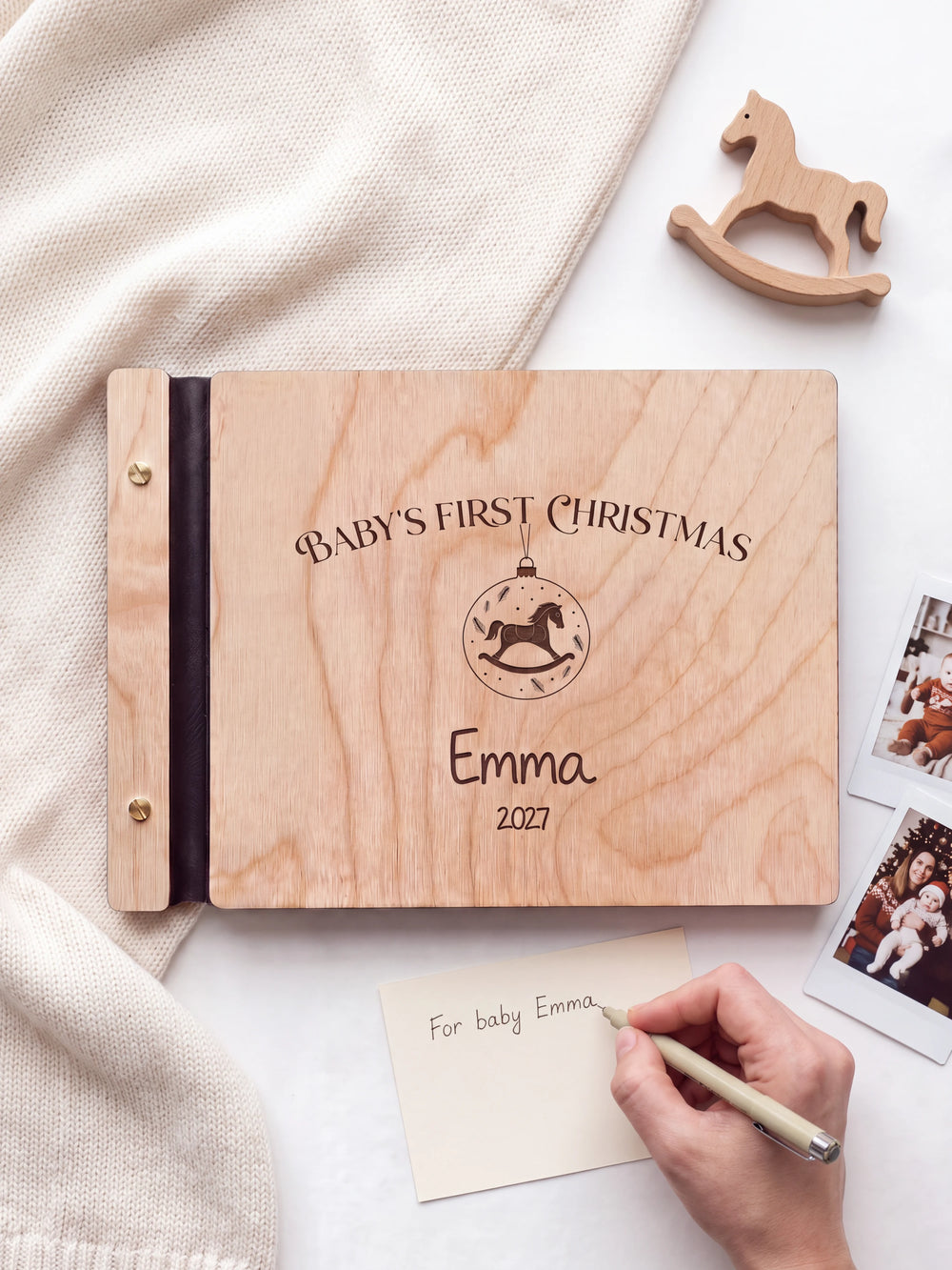 A hand writing a small gift card beside the closed Natural wood Baby's First Christmas photo book; the engraved cover and silver screws are visible