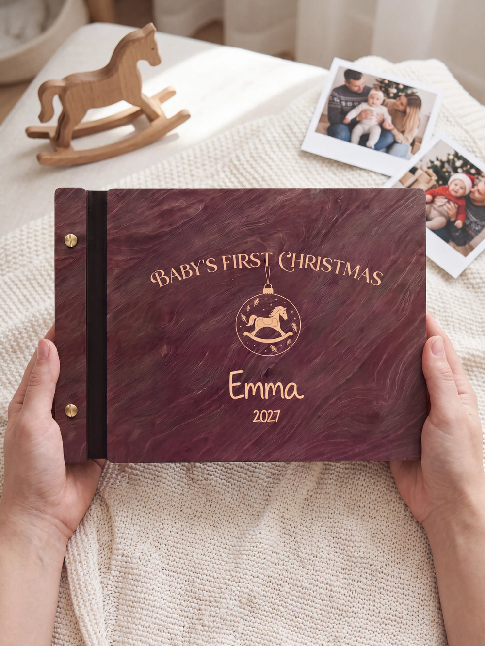 Two hands presenting the closed Jacobean Baby's First Christmas photo book with its cream engraving of a hanging ornament with a rocking horse inside, a tiny wooden rocking horse beside them