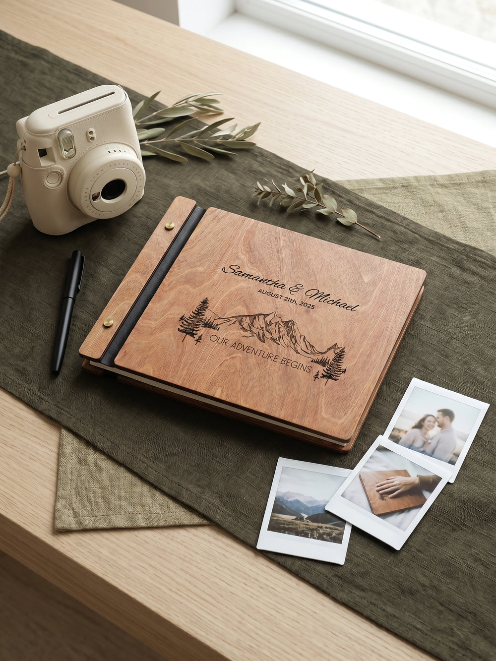 Hazelnut wood Our Adventure Begins photo guest book engraved Samantha & Michael August 21st, 2025 with mountains and pine trees, on a table with an instant camera and photos
