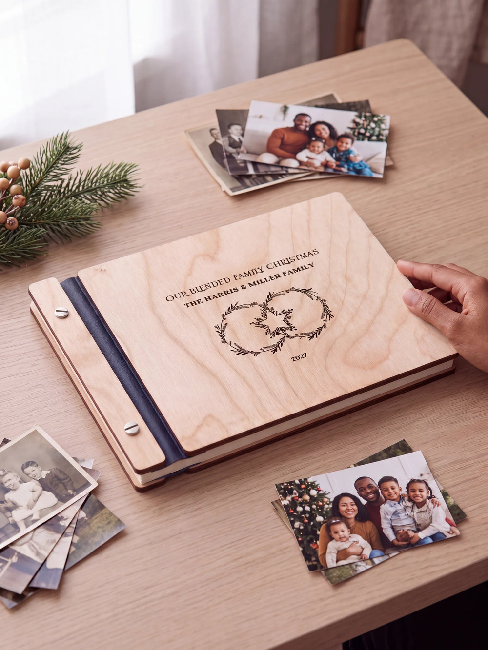 Natural wooden album with brown laser engraving of two intertwined festive wreaths, shown in a close lifestyle view with navy blue binding and silver screws. The cover reads OUR BLENDED FAMILY CHRISTMAS.