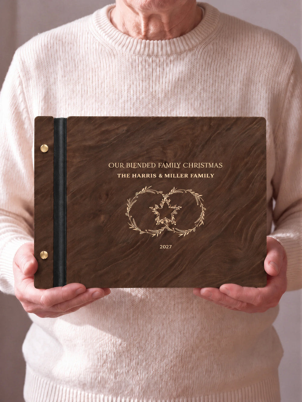 Two hands hold a Jacobean wooden photo album with the OUR BLENDED FAMILY CHRISTMAS cover facing the camera. Cream engraved artwork, the narrow binding and gold screws are visible.