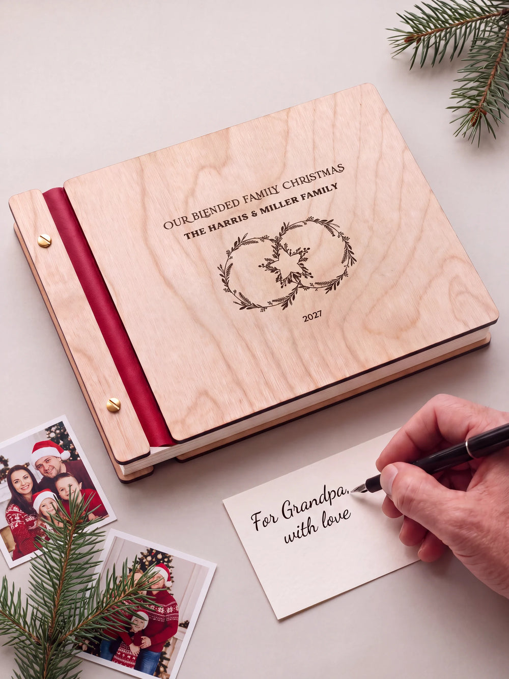 A Natural wooden album with the OUR BLENDED FAMILY CHRISTMAS engraving, cherry red binding and gold screws. A hand writes on a separate gift card beside the book.