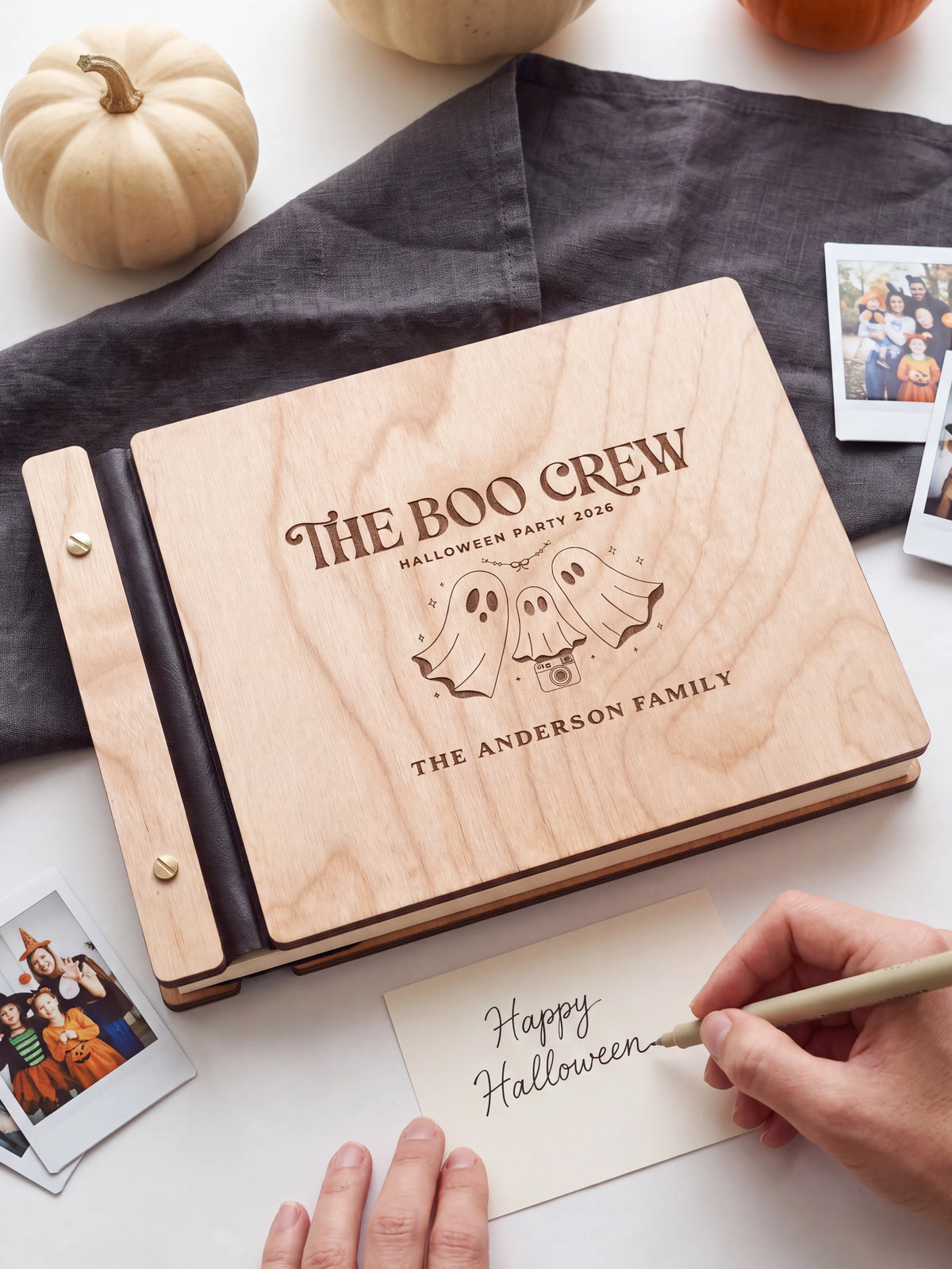 A hand writing a small gift card beside the closed Natural wood Boo Crew Halloween photo guest book; the engraved cover and silver screws are visible