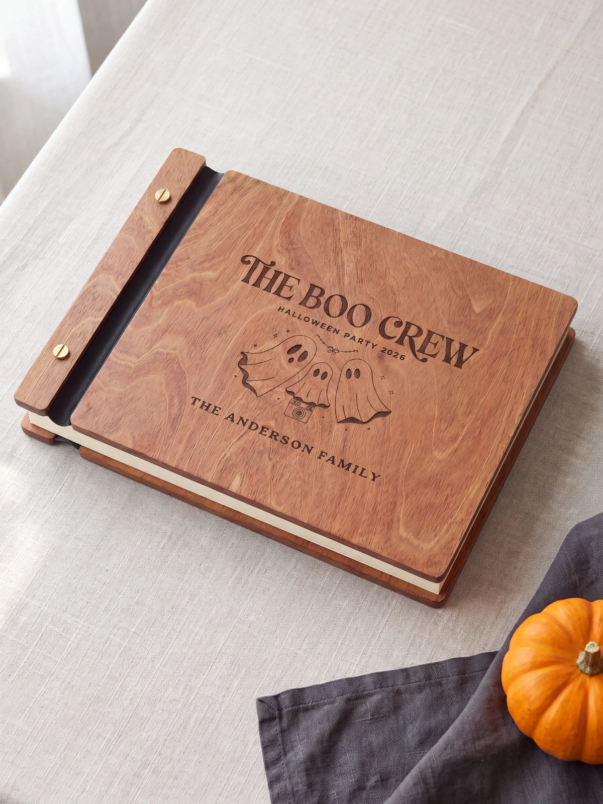 Hazelnut wood Boo Crew Halloween photo guest book lying flat on a table, the cover engraved with “THE BOO CREW”, “HALLOWEEN PARTY 2026” and “THE ANDERSON FAMILY”; two gold-tone screws along the black vegan leather spine