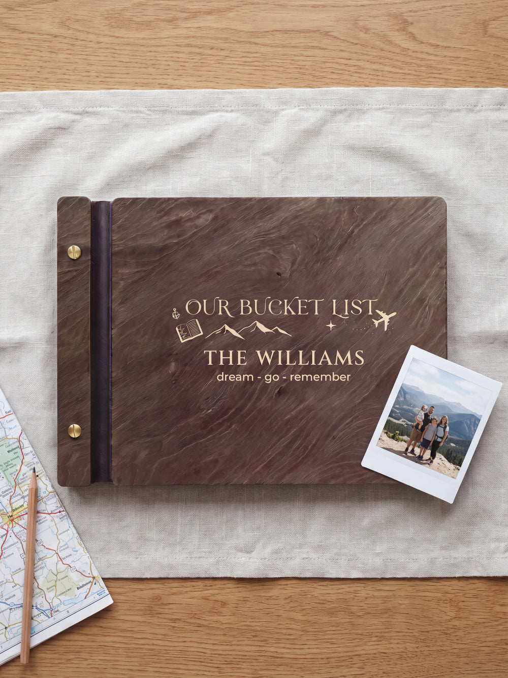 Dark brown Jacobean wooden album on a light surface, with cream engraving of mountains, a passport and an airplane and personalized cover wording. The full cover and left screw binding are visible.