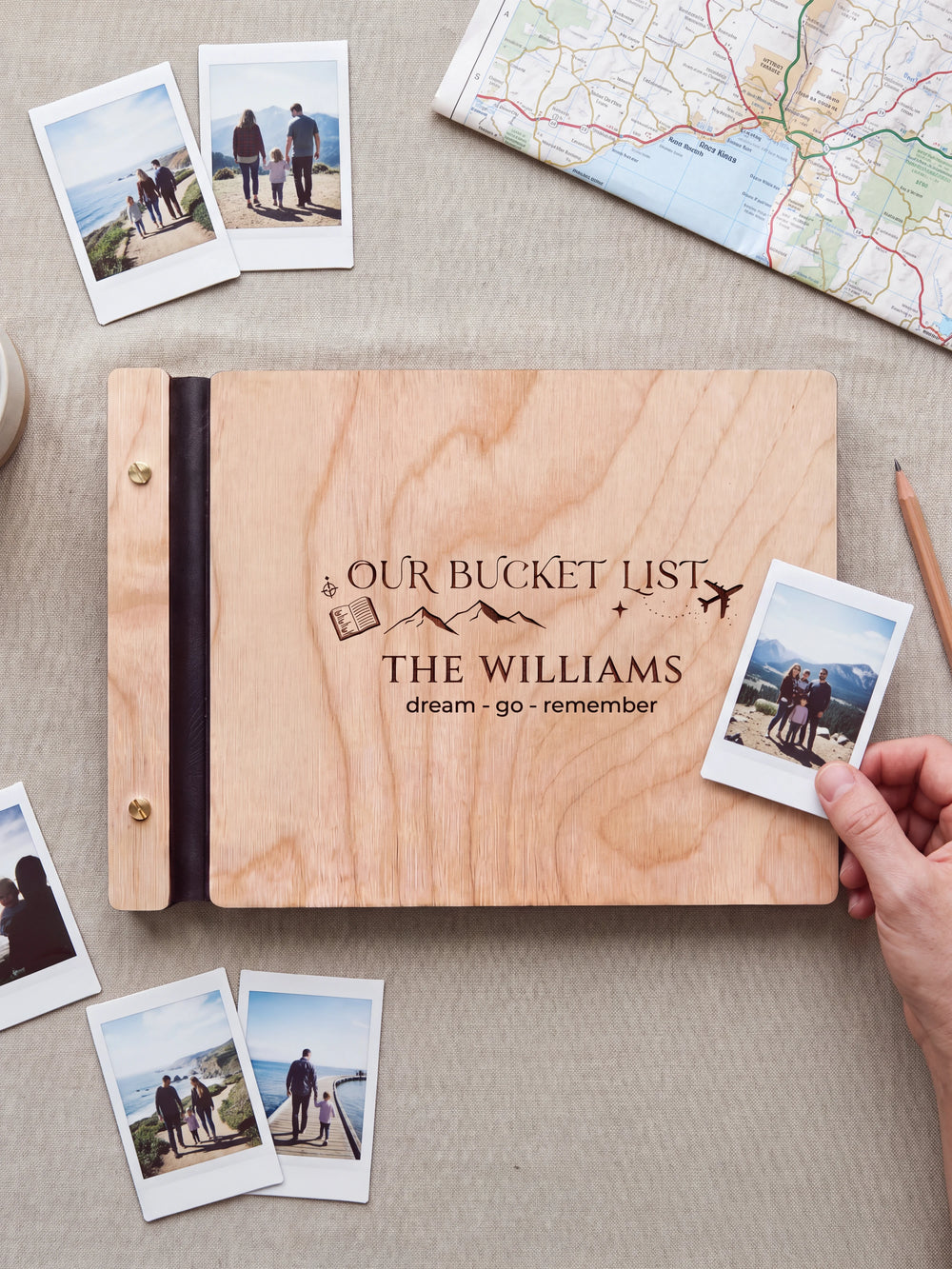 Natural wooden album with dark engraving of mountains, a passport and an airplane. A hand holds an instant photo beside the closed cover, with loose snapshots nearby.