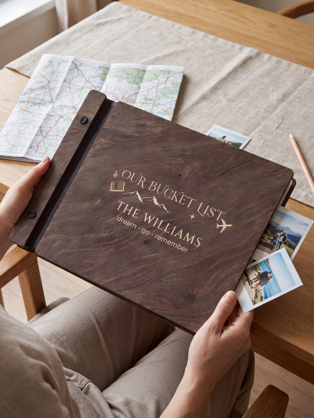 Hands hold a dark brown Jacobean wooden album close to the camera. Cream engraving shows mountains, a passport and an airplane, personalized wording and the black binding strip.