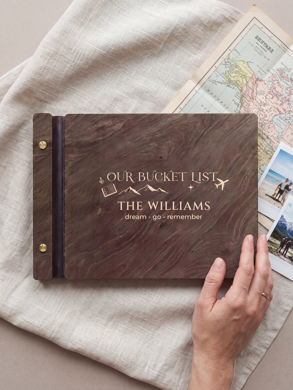 A hand rests beside the closed dark brown Jacobean album on a table. Cream engraving and personalized wording are visible, with travel photos nearby.