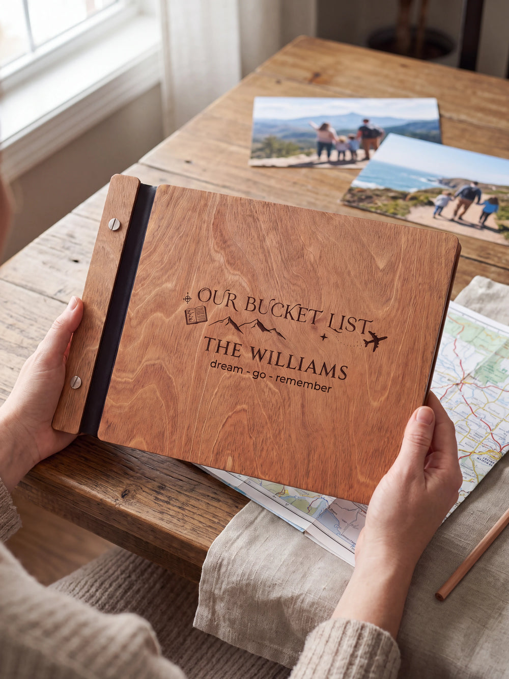 Hands hold the Hazelnut wooden album near a window. The full cover displays mountains, a passport and an airplane in dark engraving, with personalized lettering and a black binding strip.