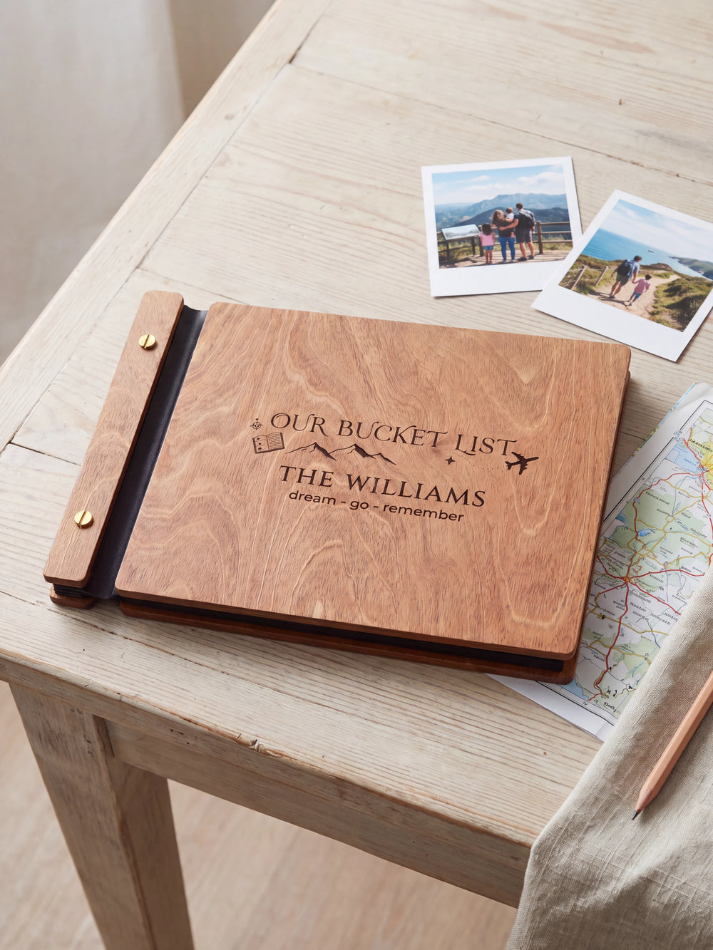 Hazelnut wooden album on a light tabletop with neutral linen. The full cover shows mountains, a passport and an airplane, personalized wording and the two-screw binding.