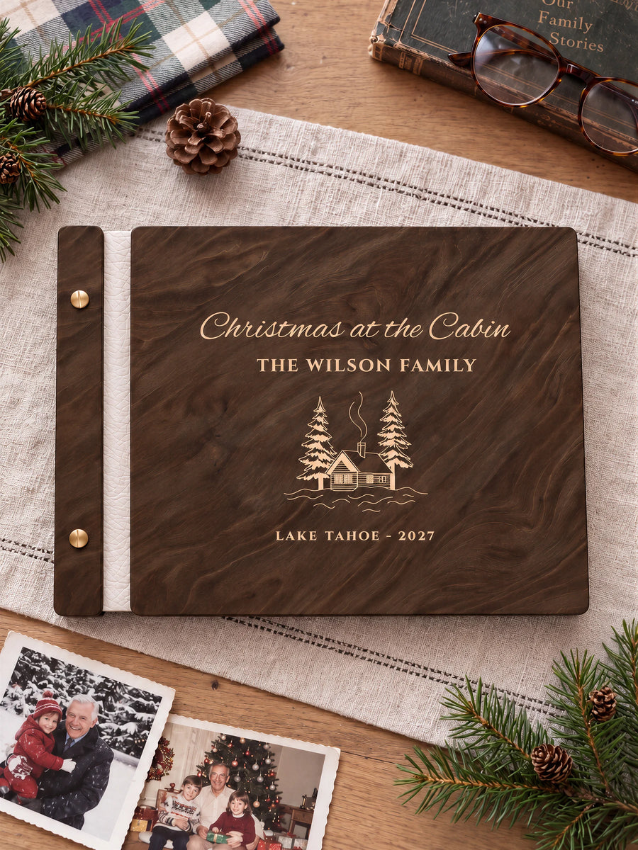 Jacobean wooden album with cream laser engraving of snowy cabin and pine trees. The full personalized cover rests on a styled surface, with ivory binding and gold screws.