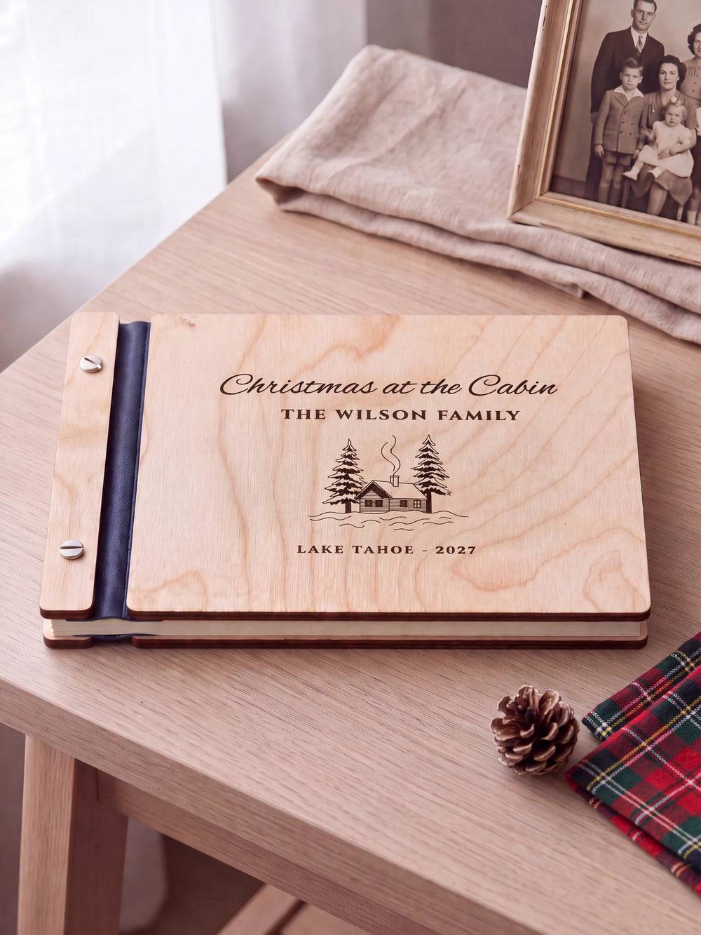 Natural wooden album with brown laser engraving of snowy cabin and pine trees, shown in a close lifestyle view with navy blue binding and silver screws. The cover reads Christmas at the Cabin.