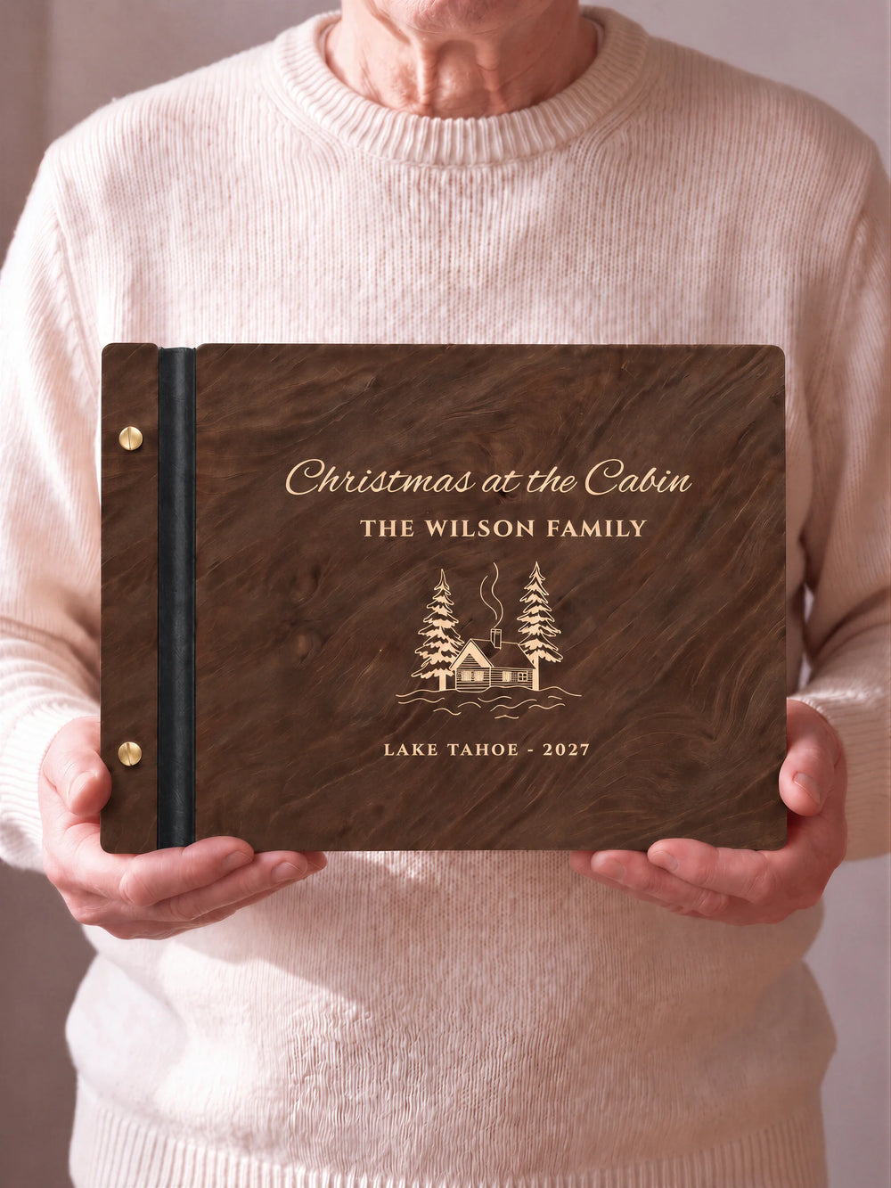 Two hands hold a Jacobean wooden photo album with the Christmas at the Cabin cover facing the camera. Cream engraved artwork, the narrow binding and gold screws are visible.