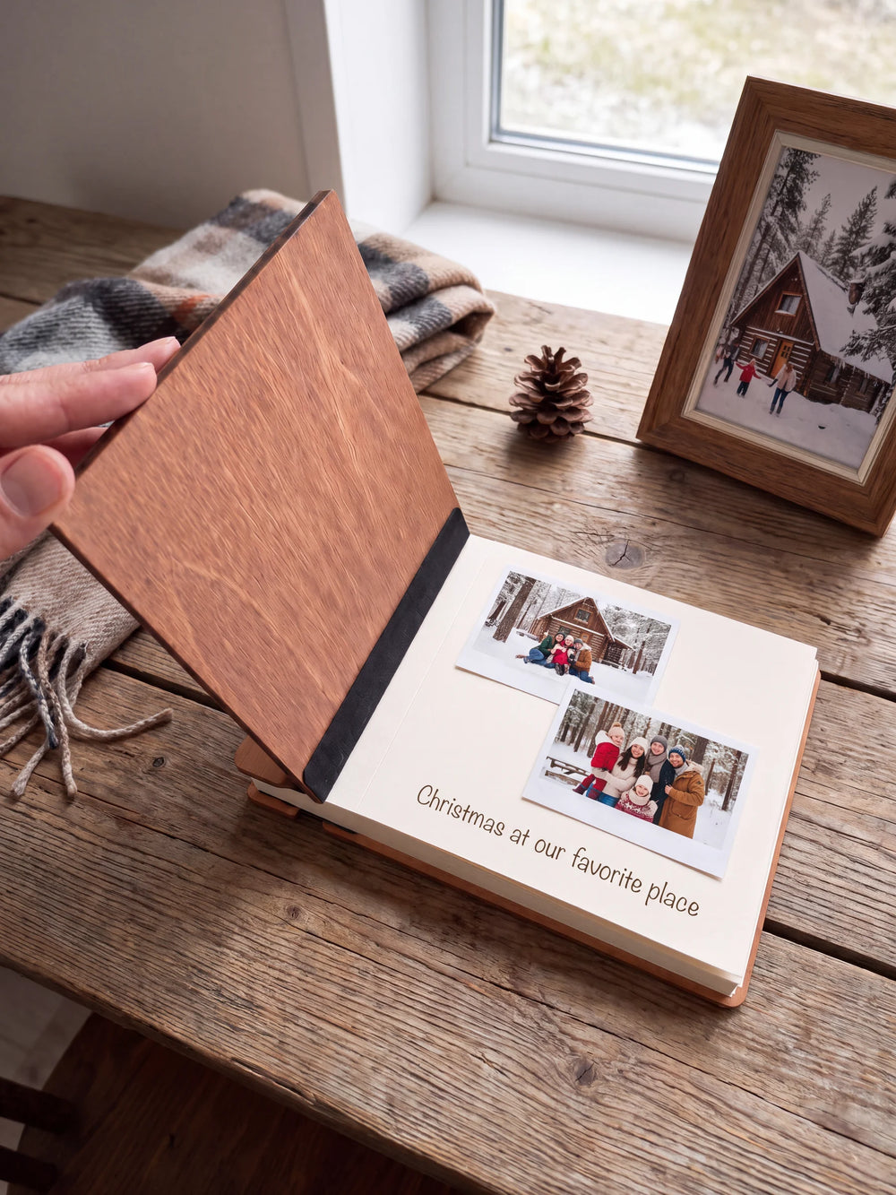 A hand lifts the Hazelnut album’s front cover, revealing a flat black inner fabric strip and a cream page arranged with instant photographs and a handwritten memory.