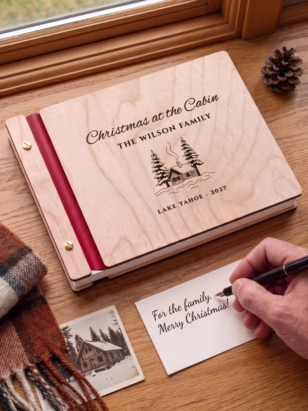 A Natural wooden album with the Christmas at the Cabin engraving, cherry red binding and gold screws. A hand writes on a separate gift card beside the book.