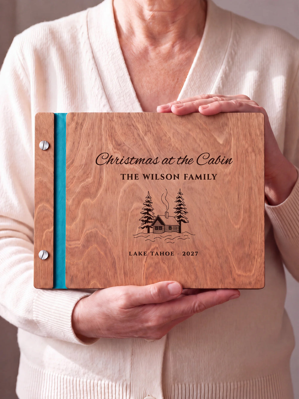 Two hands hold a Hazelnut wooden photo album. The Christmas at the Cabin cover design has brown engraving, turquoise binding and silver screws.