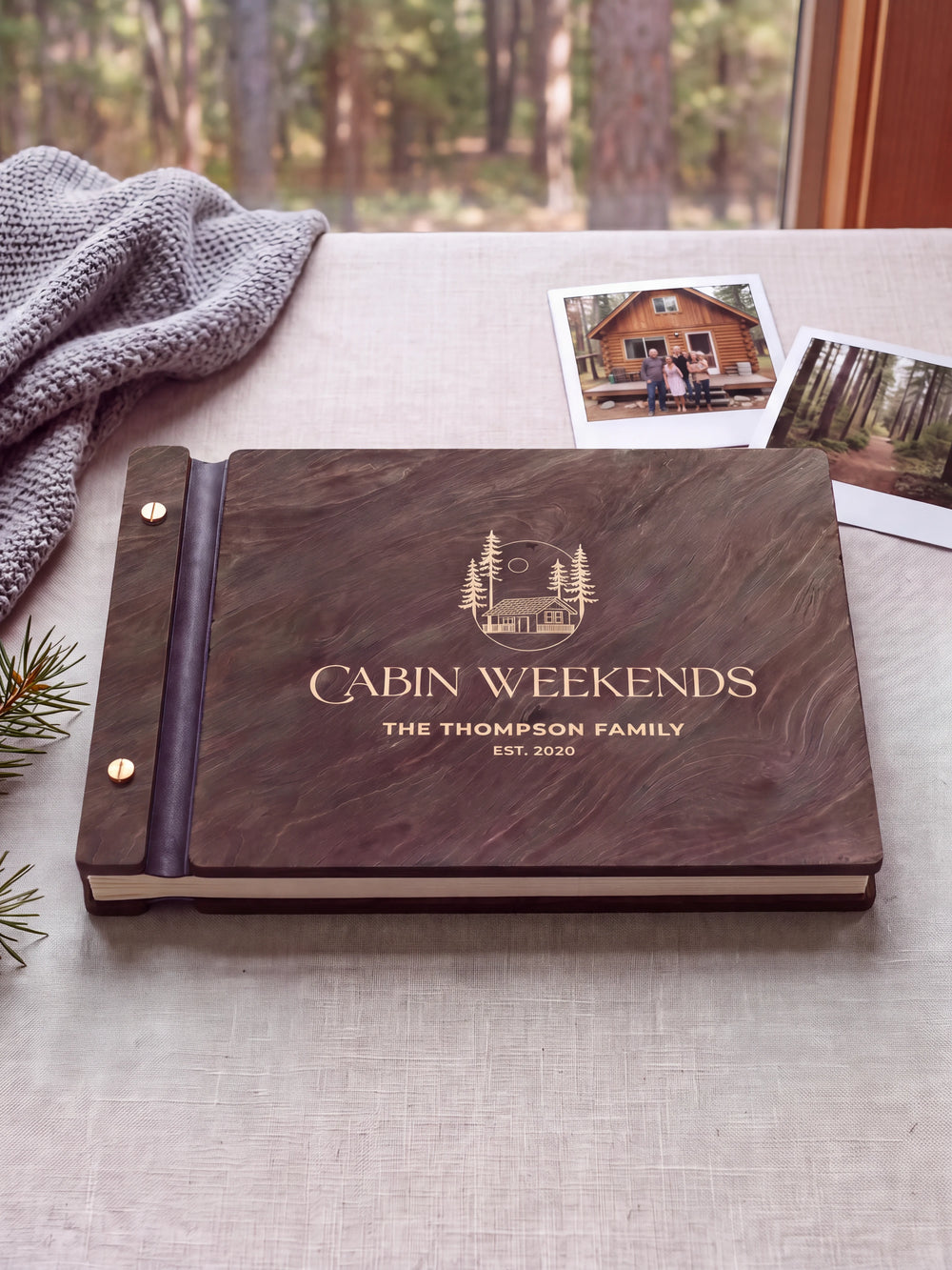 Dark brown Jacobean album on neutral linen by a cabin window, with cream engraving of a woodland cabin and pine trees. The full personalized cover, two screws and exposed cream page edges are visible.