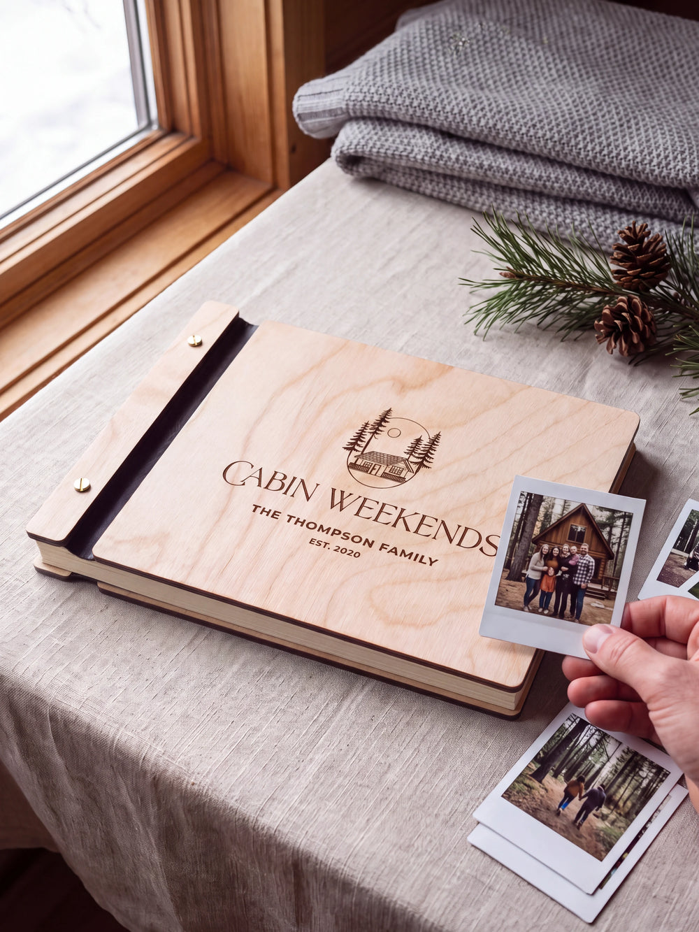 Natural wooden album with dark engraving of a woodland cabin and pine trees. A hand holds an instant photo beside the closed cover, with loose snapshots nearby.