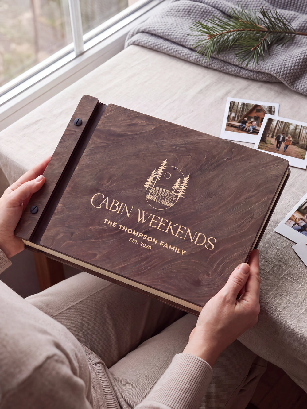 Hands hold a dark brown Jacobean wooden album close to the camera. Cream engraving shows a woodland cabin and pine trees, personalized wording and the black binding strip.