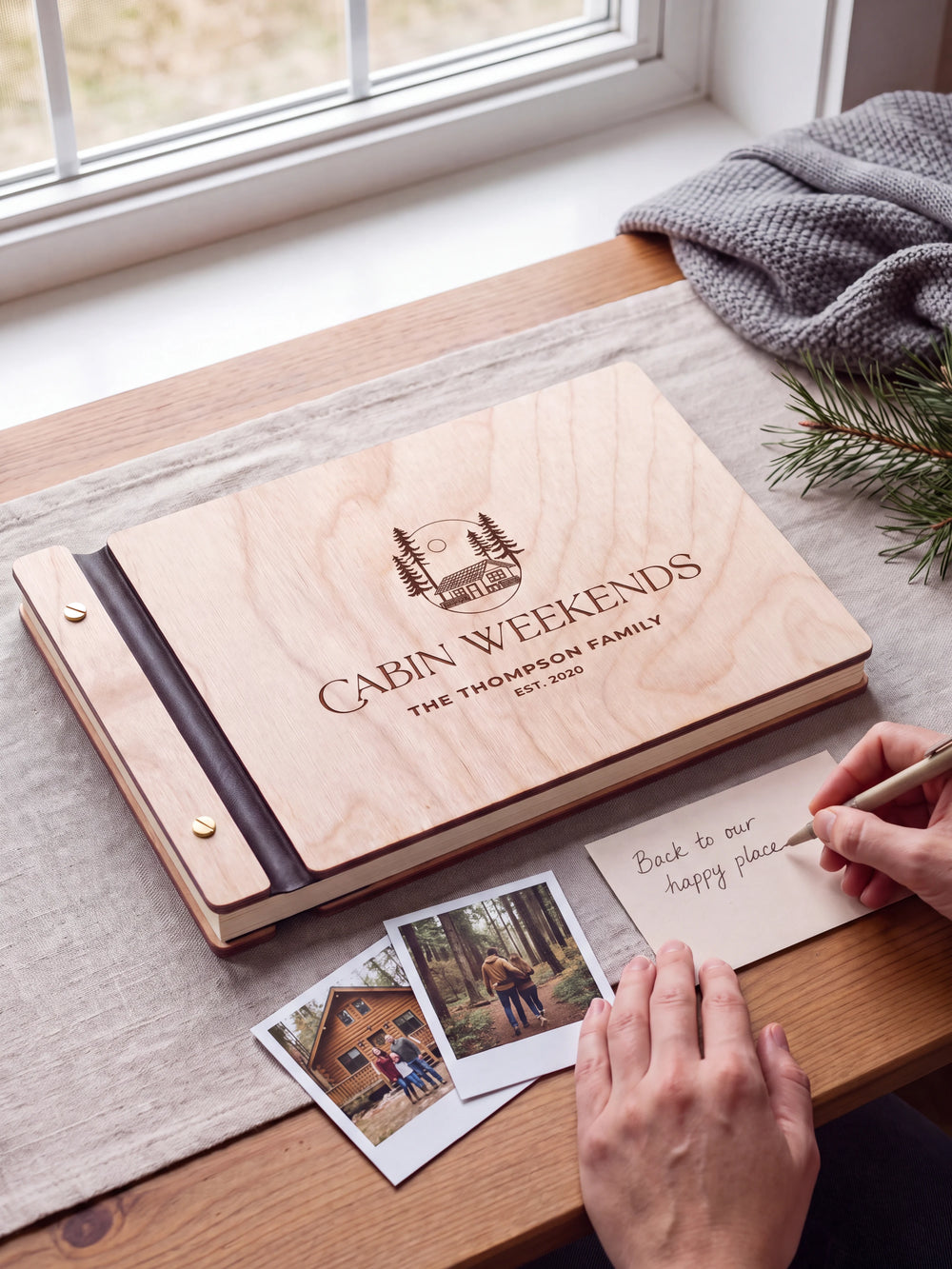 A Natural wooden album showing a woodland cabin and pine trees rests beside a separate gift card. A hand writes 