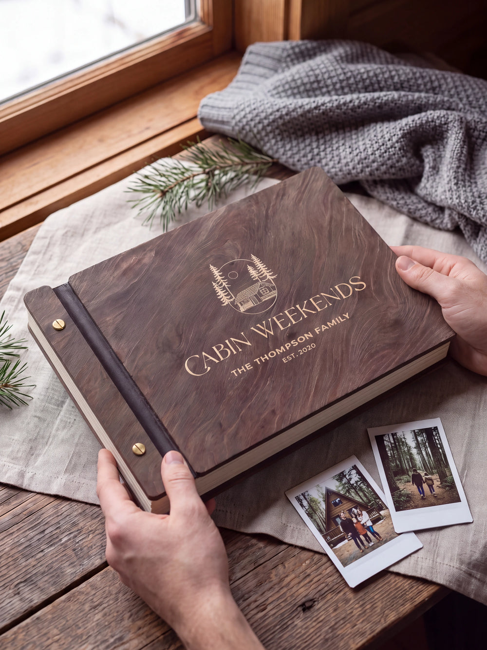 Two hands present a dark brown Jacobean wooden album, with the full cream-engraved woodland cabin and pine trees design and personalized cover wording facing the camera.
