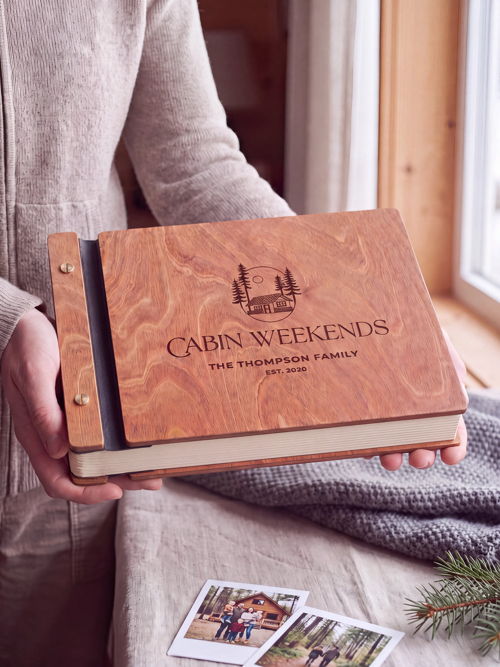 Hands hold the Hazelnut wooden album near a window. The full cover displays a woodland cabin and pine trees in dark engraving, with personalized lettering and a black binding strip.