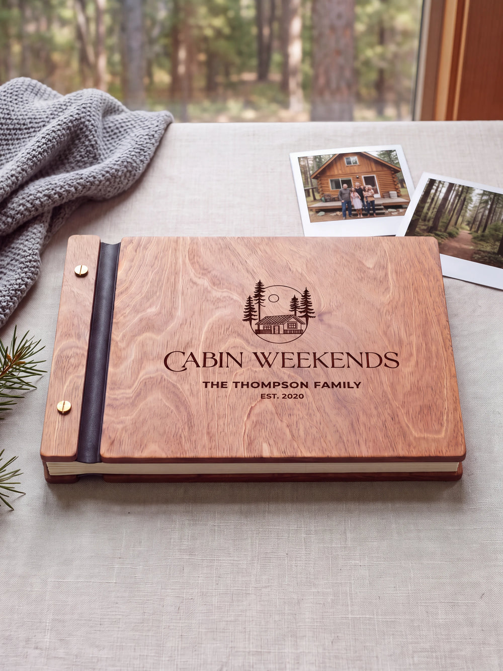 Hazelnut wooden album on a light tabletop with neutral linen. The full cover shows a woodland cabin and pine trees, personalized wording and the two-screw binding.