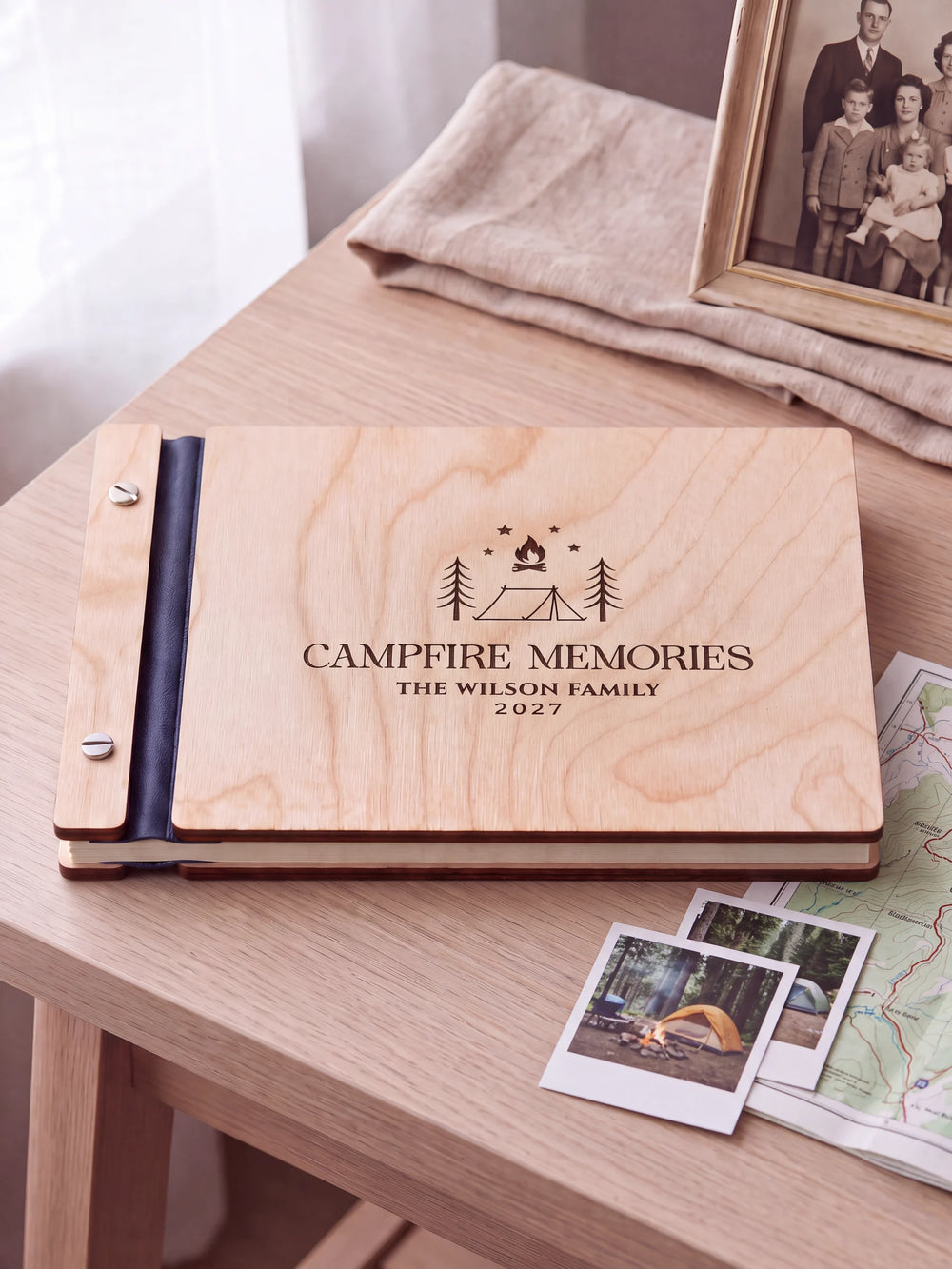 Natural wooden album with brown laser engraving of tent, campfire, pine trees and stars, shown in a close lifestyle view with navy blue binding and silver screws. The cover reads CAMPFIRE MEMORIES.