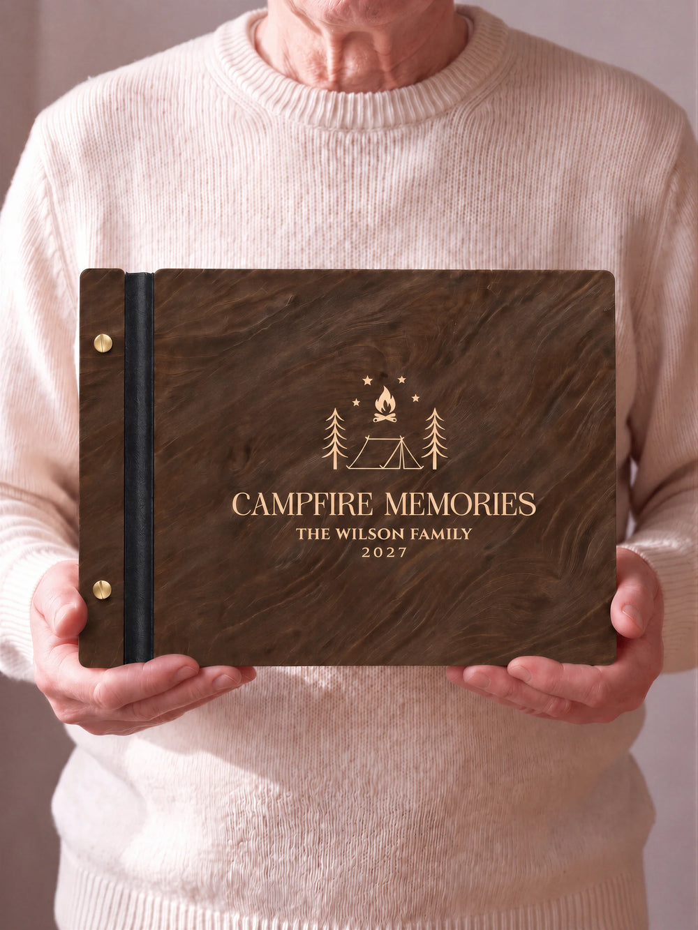 Two hands hold a Jacobean wooden photo album with the CAMPFIRE MEMORIES cover facing the camera. Cream engraved artwork, the narrow binding and gold screws are visible.