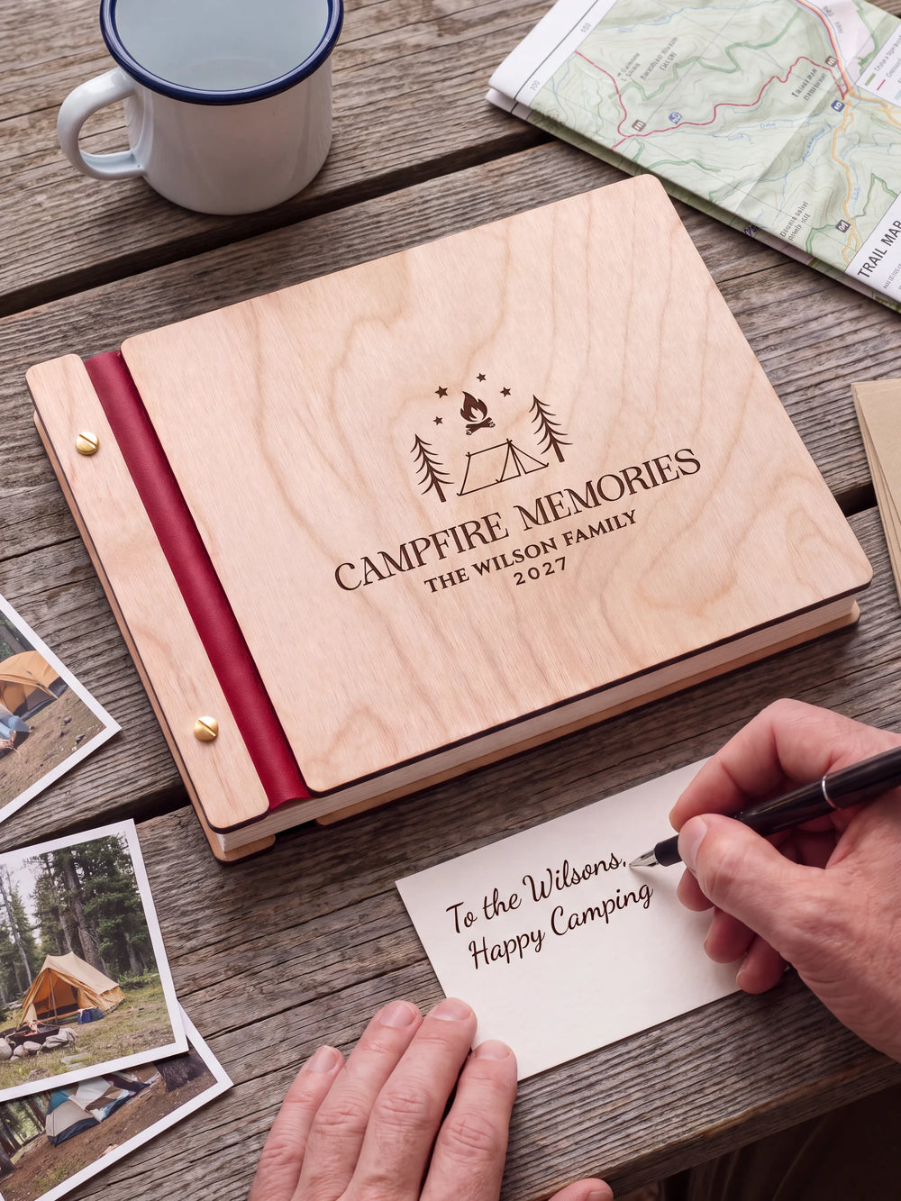 A Natural wooden album with the CAMPFIRE MEMORIES engraving, cherry red binding and gold screws. A hand writes on a separate gift card beside the book.