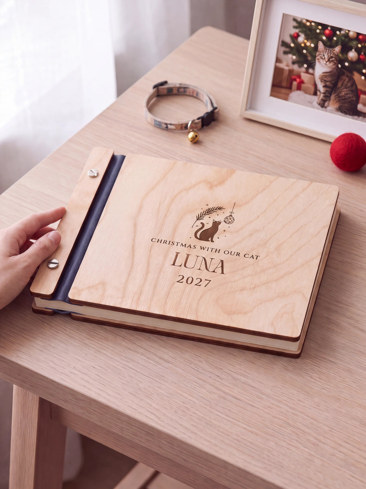 Natural wooden album with brown laser engraving of cat silhouette, hanging ornament and foliage, shown in a close lifestyle view with navy blue binding and silver screws. The cover reads CHRISTMAS WITH OUR CAT.