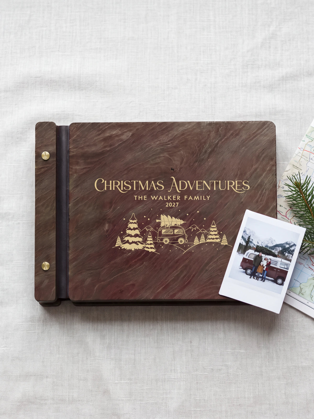 Jacobean wood Christmas Adventures photo book lying flat on a cabin coffee table, the cover engraved with “CHRISTMAS ADVENTURES”, “THE WALKER FAMILY” and “2027”; two gold-tone screws along the black vegan leather spine