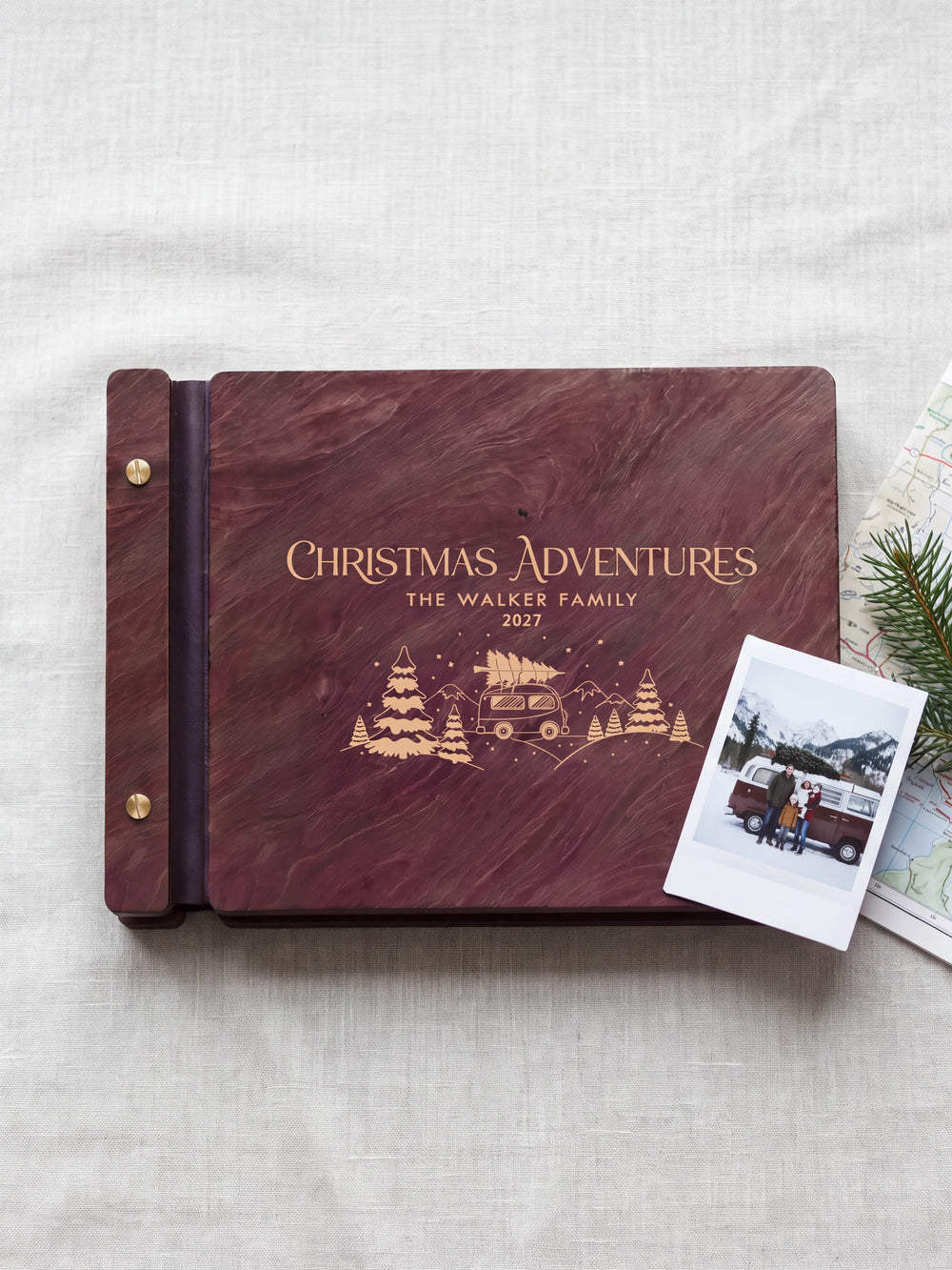 Jacobean wood Christmas Adventures photo book lying flat on a cabin coffee table, the cover engraved with “CHRISTMAS ADVENTURES”, “THE WALKER FAMILY” and “2027”; two gold-tone screws along the black vegan leather spine