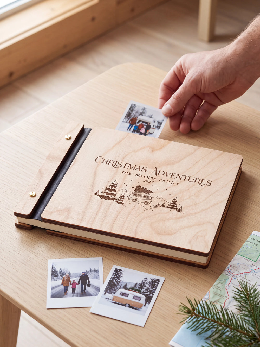 Natural wood Christmas Adventures photo book on a light table while a hand picks an instant photo to add; the engraved cover reads “CHRISTMAS ADVENTURES”, “THE WALKER FAMILY” and “2027”