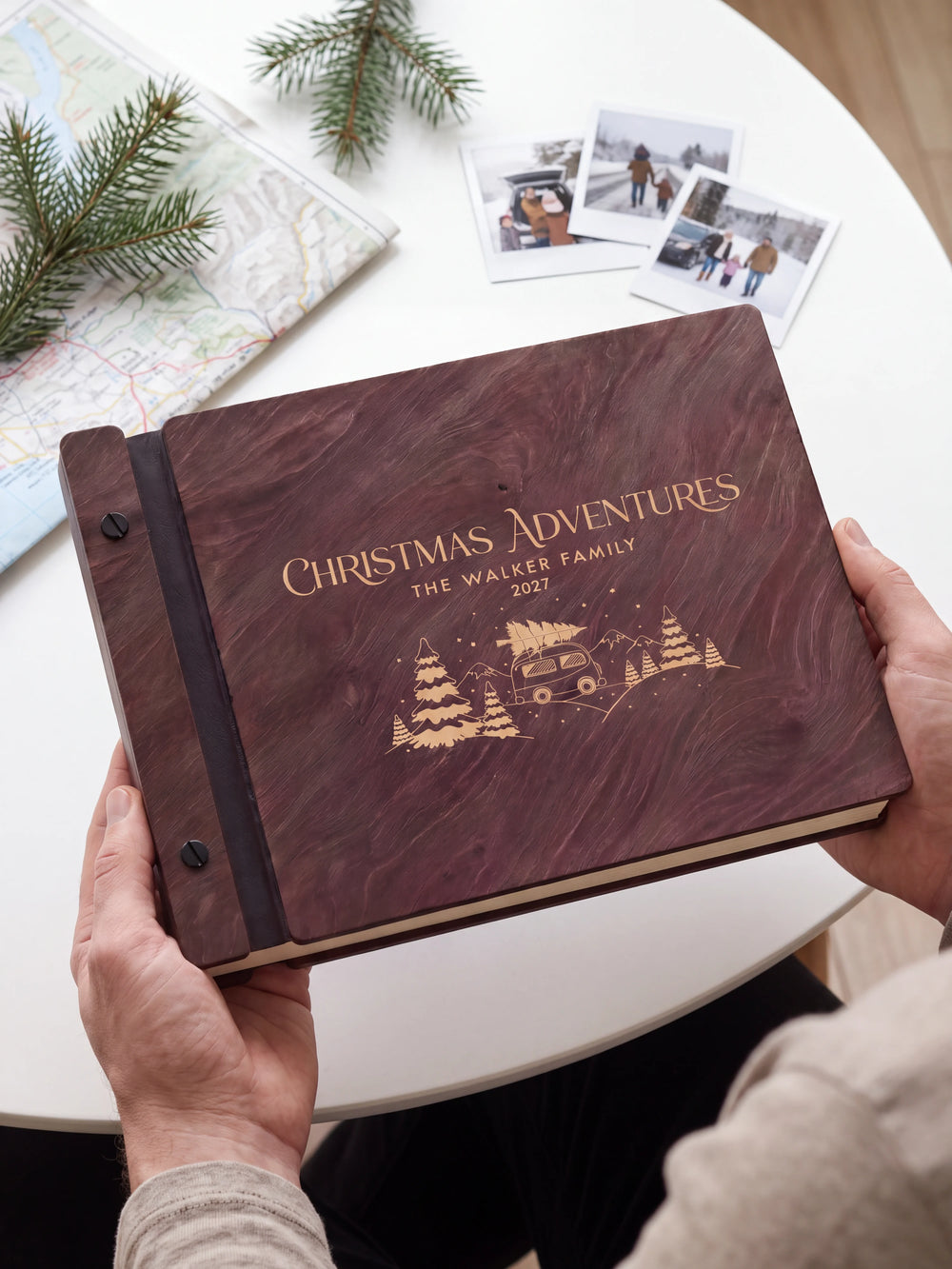Two hands holding the Jacobean Christmas Adventures photo book up to the camera, the cream engraving of a camper van carrying a Christmas tree among snowy pines and “CHRISTMAS ADVENTURES”, “THE WALKER FAMILY” and “2027” filling the cover