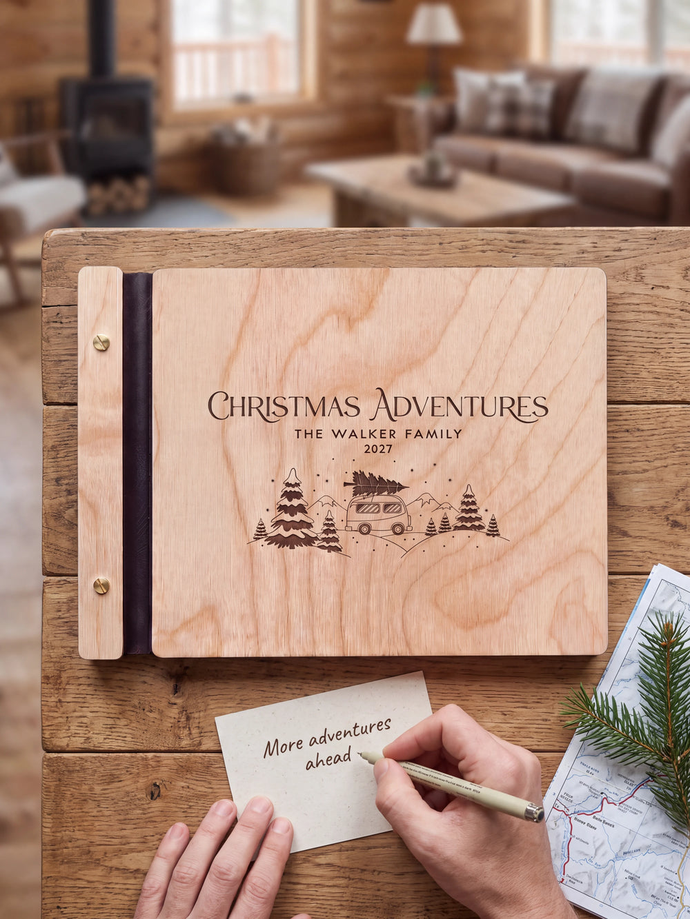 A hand writing a small gift card beside the closed Natural wood Christmas Adventures photo book; the engraved cover and silver screws are visible