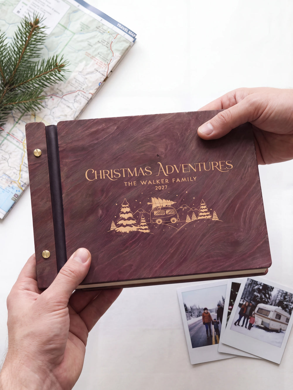 Two hands presenting the closed Jacobean Christmas Adventures photo book with its cream engraving of a camper van carrying a Christmas tree among snowy pines, a folded travel map and a pine sprig beside them