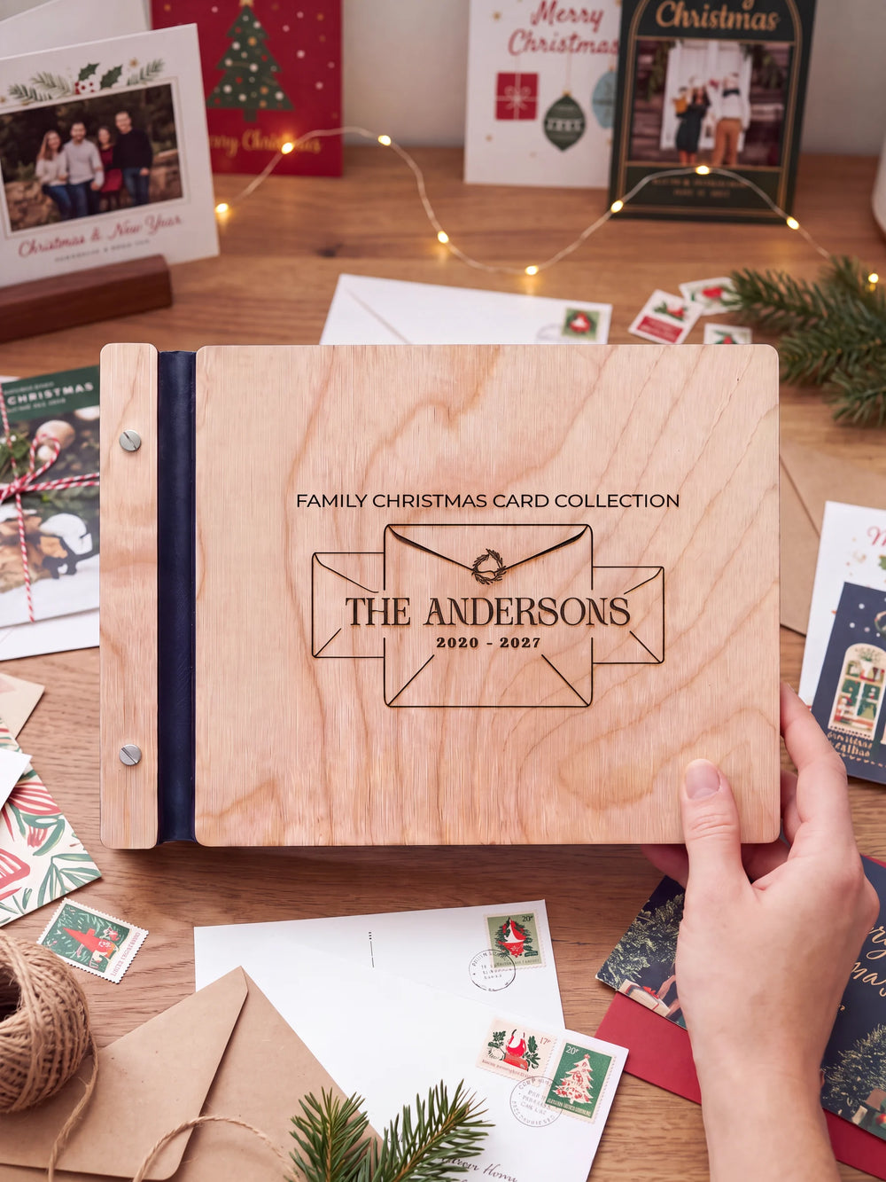 Natural wooden album with brown laser engraving of three envelopes and a tiny wreath, shown in a close lifestyle view with navy blue binding and silver screws. The cover reads FAMILY CHRISTMAS CARD COLLECTION.