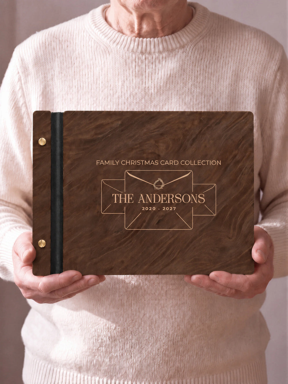Two hands hold a Jacobean wooden photo album with the FAMILY CHRISTMAS CARD COLLECTION cover facing the camera. Cream engraved artwork, the narrow binding and gold screws are visible.