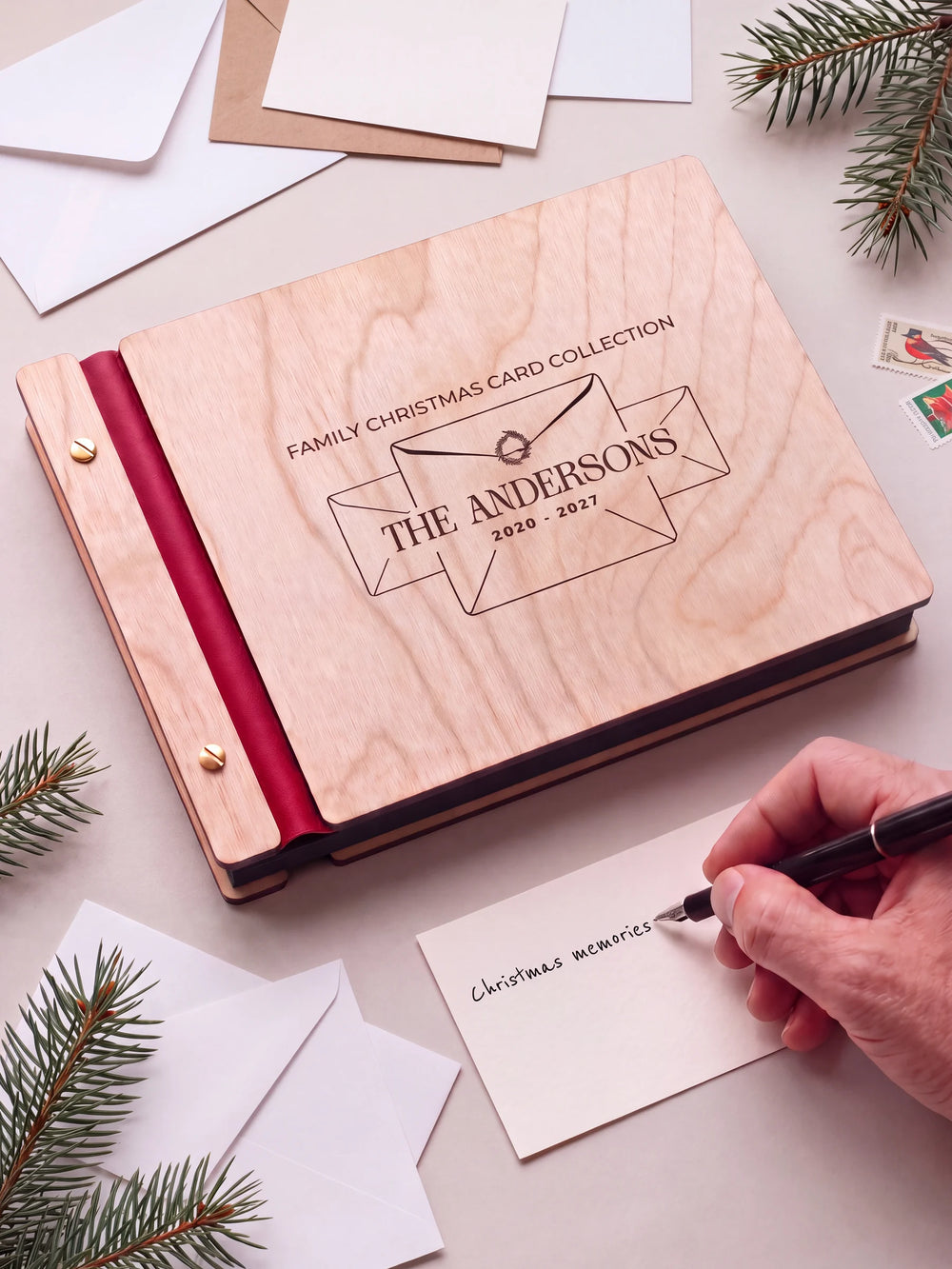 A Natural wooden album with the FAMILY CHRISTMAS CARD COLLECTION engraving, cherry red binding and gold screws. A hand writes on a separate gift card beside the book.