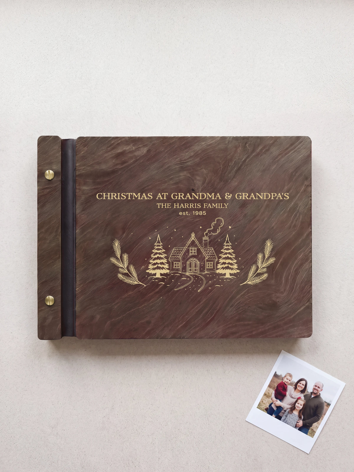 Dark Jacobean wooden album on pale fabric, with gold-tone engraving of a snowy cottage and the words Christmas at Grandma & Grandpa's, The Harris Family, est. 1985; two gold-tone screws line the left edge